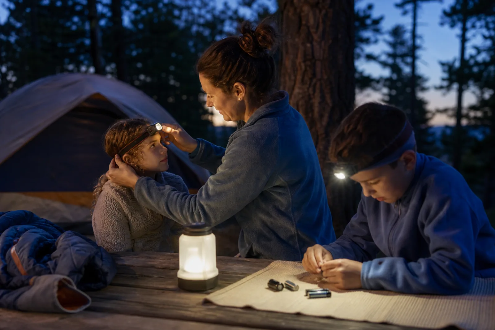 A parent adjusts a child's headlamp while another child works by lantern light.