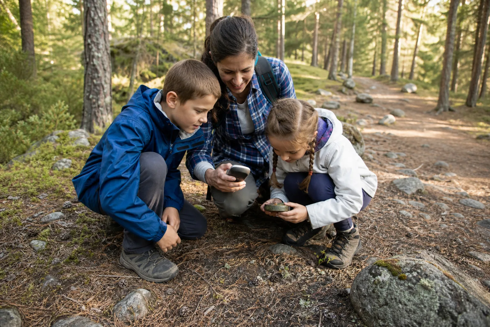 A parent supervises two children examining a small geocache beside a forest trail.