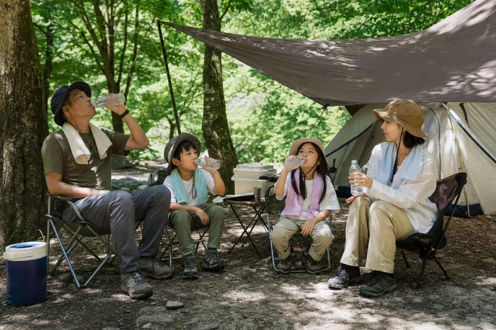 A family rests and drinks water together beneath a shaded campsite tarp.