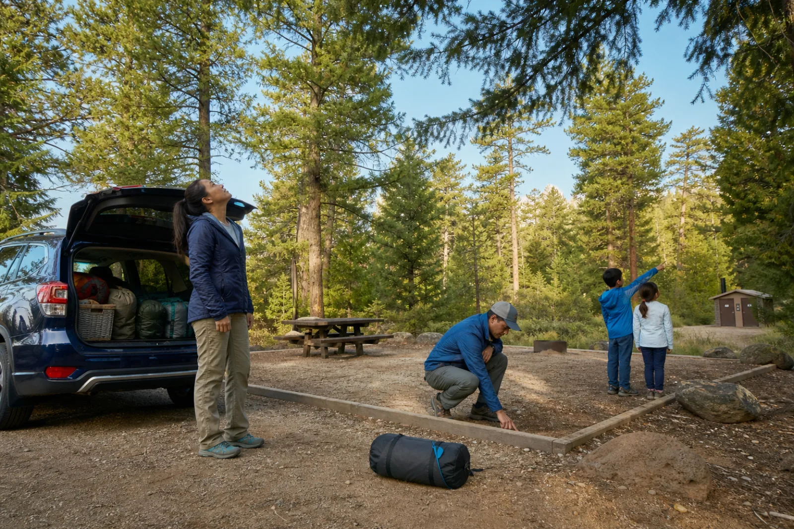 A family checks trees, drainage, and paths before unpacking a forest campsite.